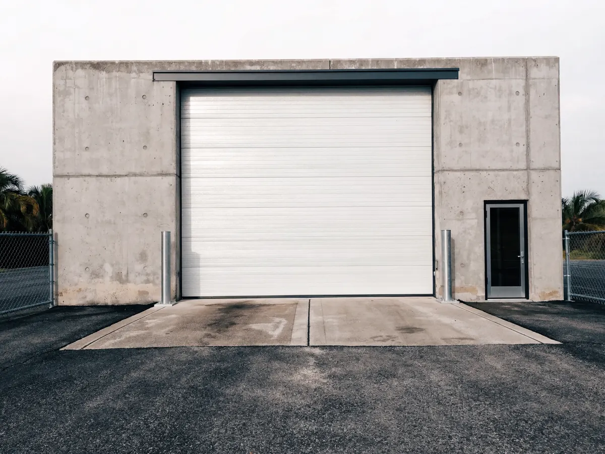 Wide ribbed white steel commercial sectional door in a concrete tilt wall unit of a Florida industrial park, with a loading apron and a bollard either side