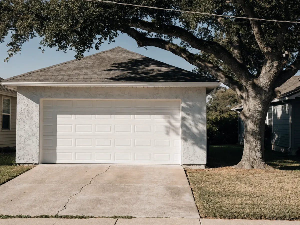 White vinyl garage door with a smooth flush surface and long shallow raised panels, on a modest single storey stucco house under a live oak in an older inland neighbourhood