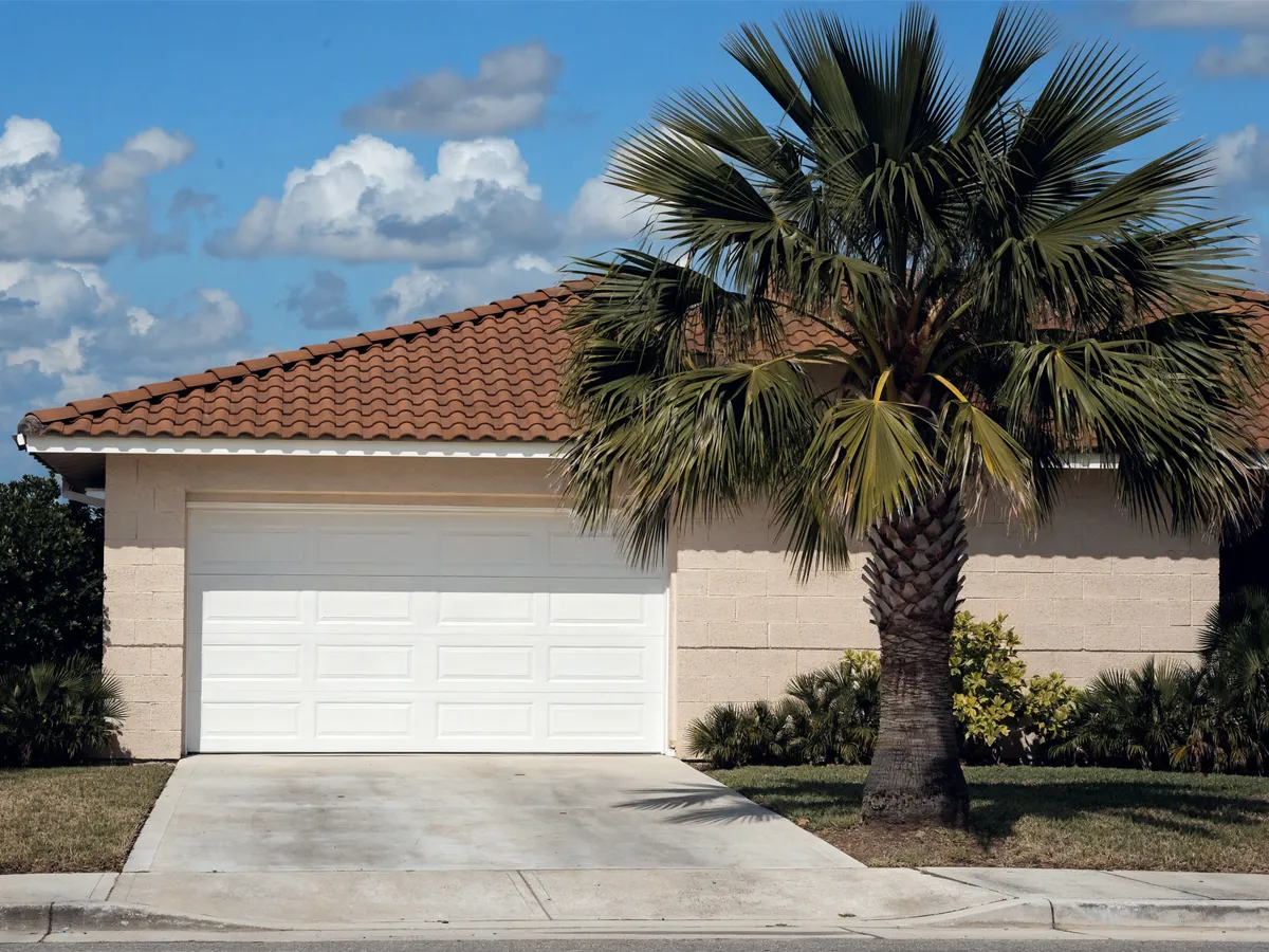 Single sixteen foot white steel double garage door on a 1980s concrete block and stucco house with a barrel tile roof and a palm in the yard