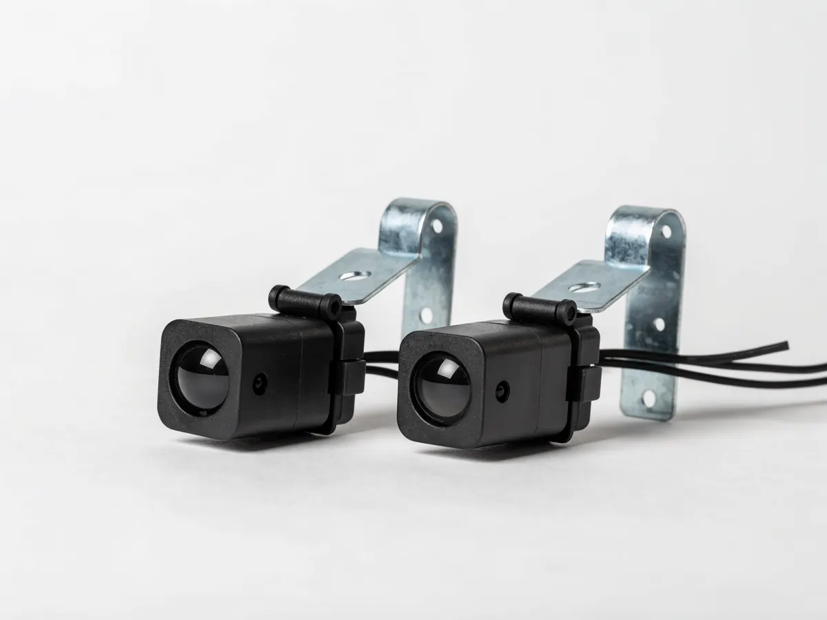 Pair of garage door photo eye safety sensors in adjustable steel brackets, each with a round lens and an indicator light, thin bell wire trailing from the back